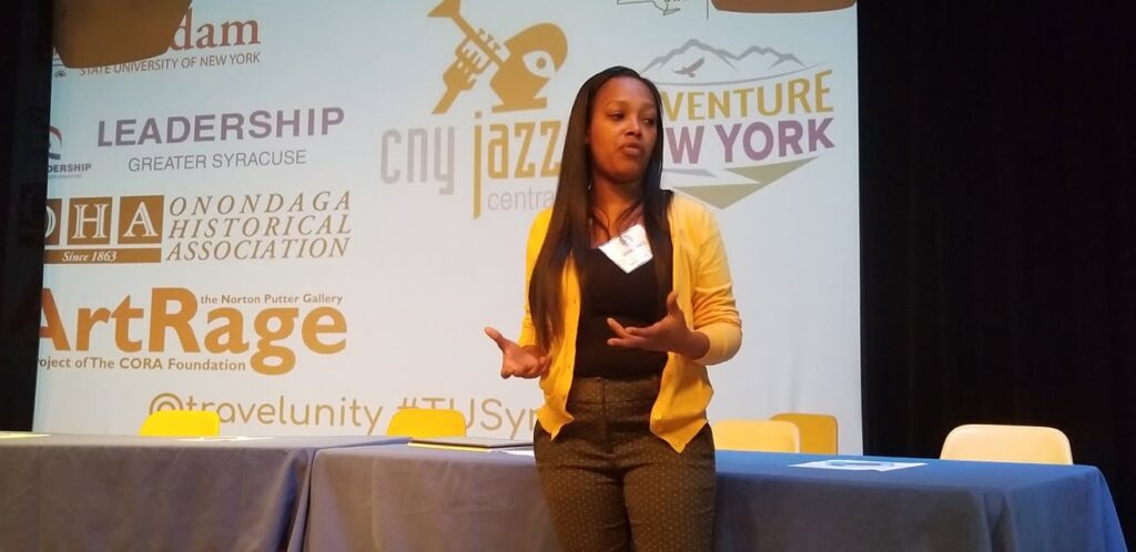 Speaker presenting on stage at the 2019 Travel Unity Summit in Syracuse, New York, with sponsor and community partner logos displayed on the backdrop.