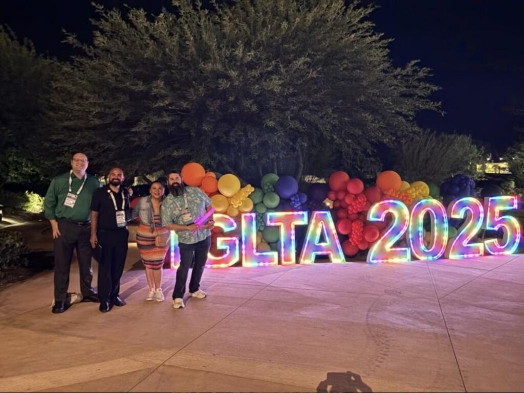 Convention attendees wearing name badges stand beside a lit IGLTA 2025 sign and rainbow balloon display at the IGLTA Global Convention in Palm Springs.
