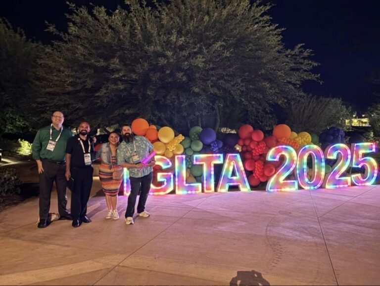Convention attendees wearing name badges stand beside a lit IGLTA 2025 sign and rainbow balloon display at the IGLTA Global Convention in Palm Springs.